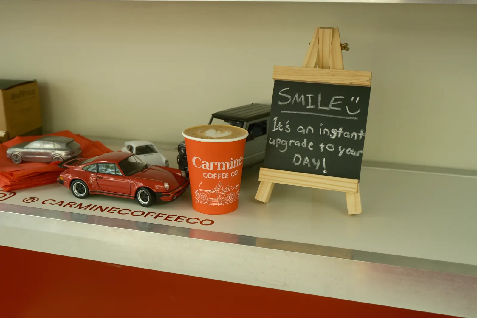 A Carmine cup with latte art on a counter, beside a red model Porsche and a chalkboard that reads Smile